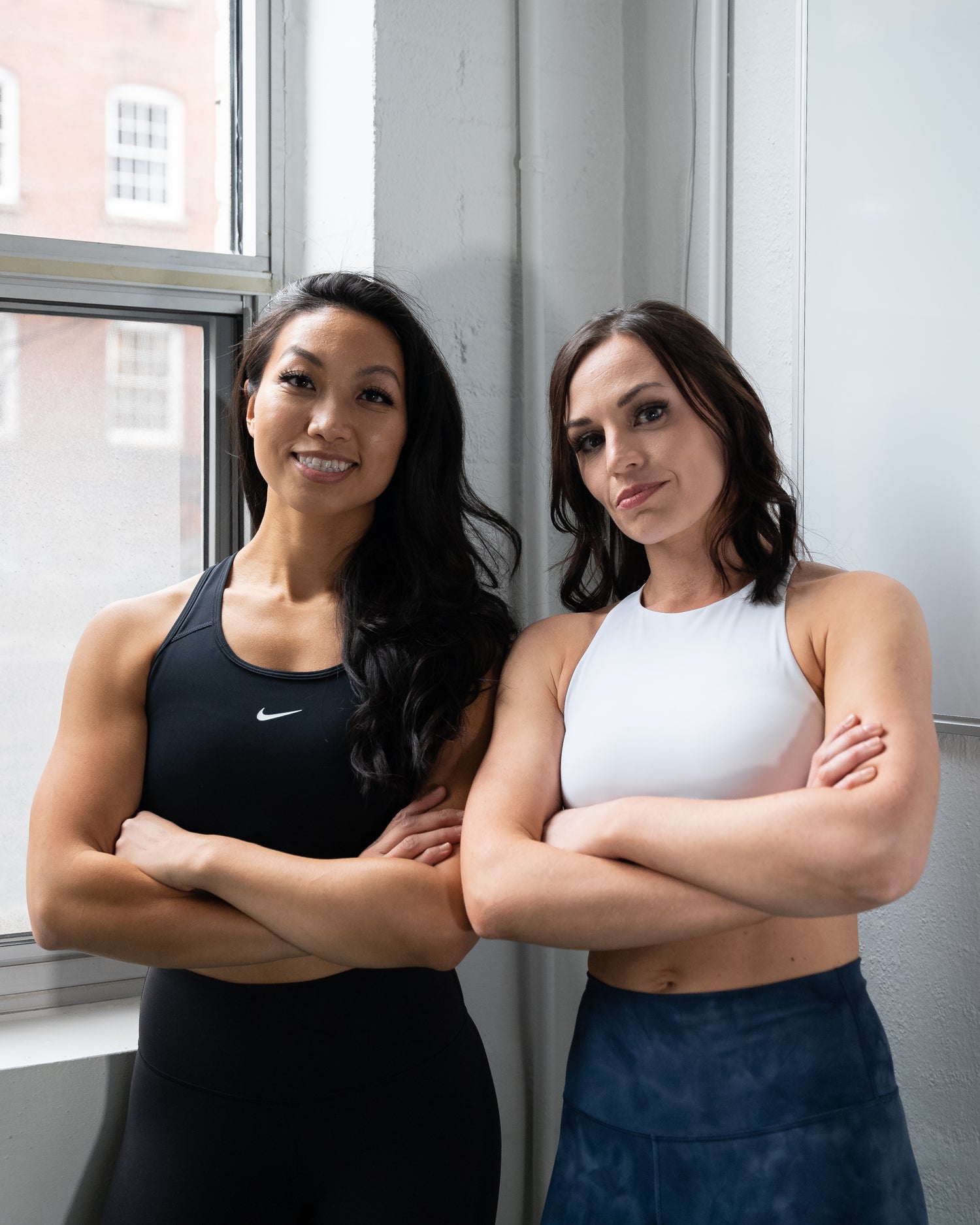 Two female personal trainers standing by a window with arms crossed. Personal trainer one is wearing a black sports bra and leggings. Personal trainer two is wearing a white sports bra and blue leggings.